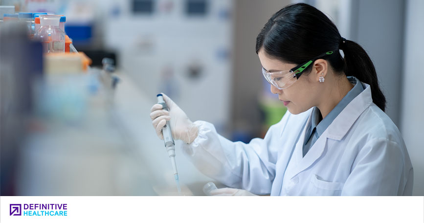 A female researcher in a lab coat and safety glasses uses a handheld instrument in a lab setting.