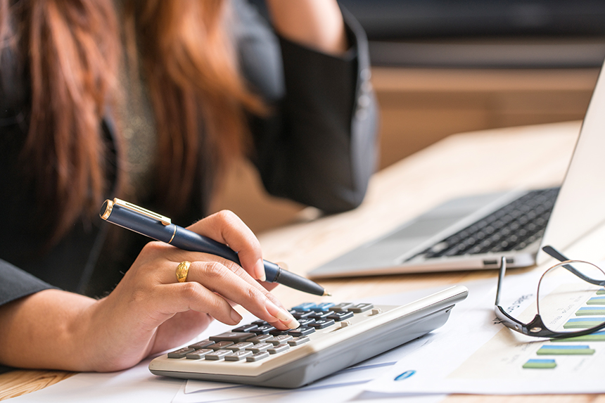 A closeup of a desk and a hand holding a pen as it types on a calculator. 