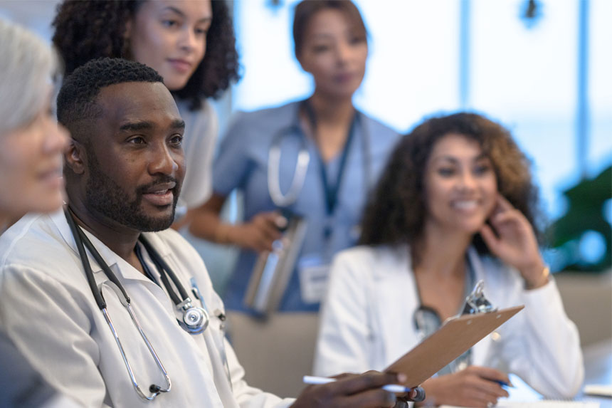 Several smiling physicians sit and stand around a table and listen to a speaker (out of frame).