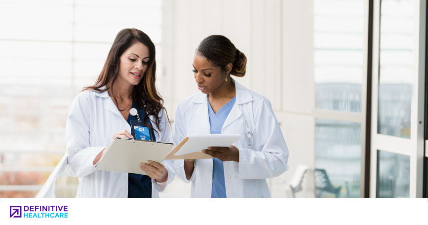 Two female healthcare professionals stand in a well-lit room and compare charts.