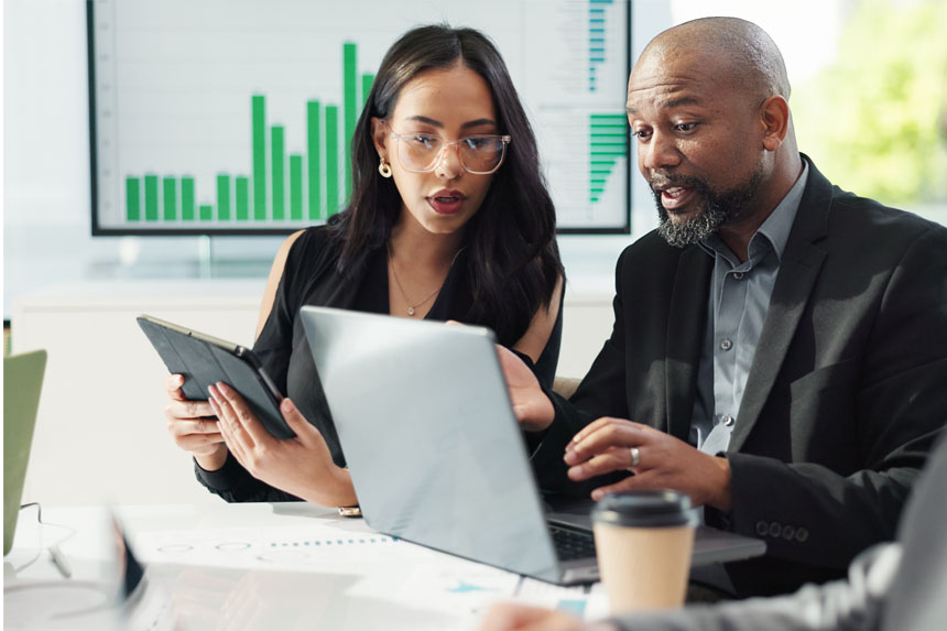 Two people in business attire sit at a table together with a chart displayed behind them. One holds a tablet as they both look at the other's laptop screen.