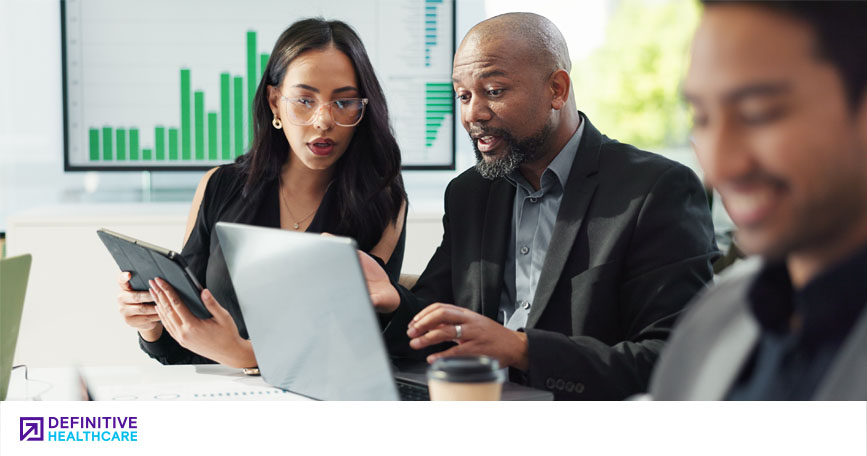 Two people in business attire sit at a table together with a chart displayed behind them. One holds a tablet as they both look at the other's laptop screen. Another person in business attire sits smiling (out of focus).
