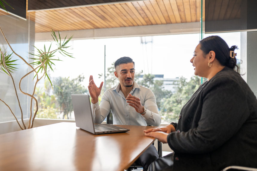 A man and woman in business casual attire sit at a table talking. There is a laptop on the table between them.