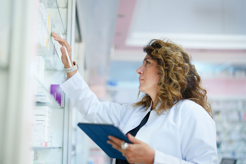 A female healthcare professional in a lab coat touches a box on a pharmacy shelf with a stylus pen in hand, and she holds a tablet in the other hand.