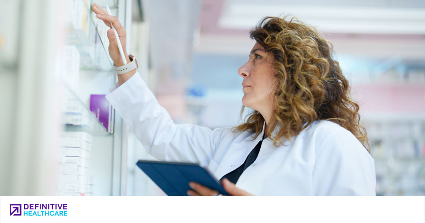 A female healthcare professional in a lab coat touches a box on a pharmacy shelf with a stylus pen in hand, and she holds a tablet in the other hand.