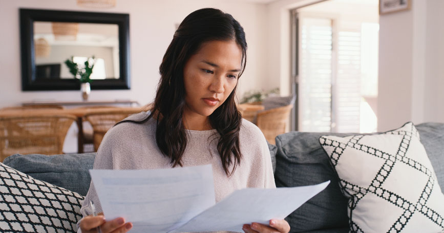 A female sits on a couch while holding up a piece of paper in each hand to read.