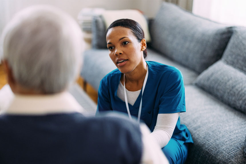 A female healthcare worker kneels down to speak with a patient who is seated.