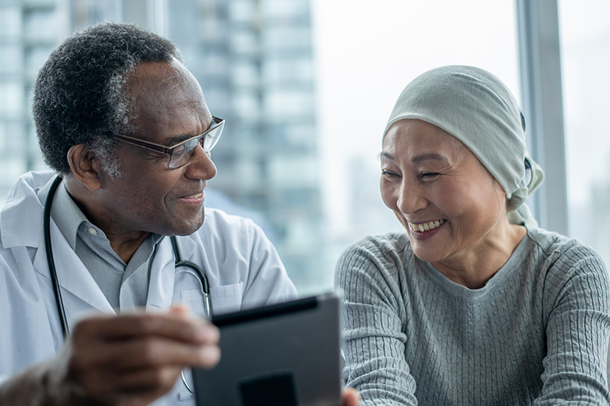 A male physician and female patient sit together smiling. The physician is holding up a tablet.