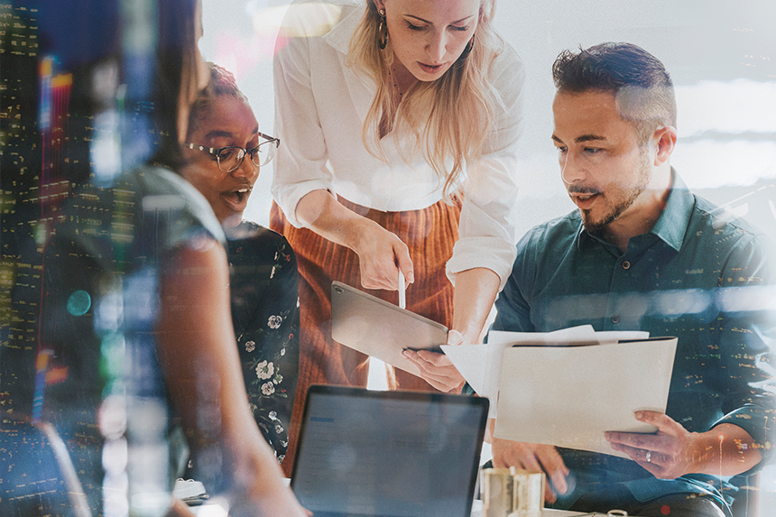 Marketing team members huddle over folders and a laptop to discuss strategy.