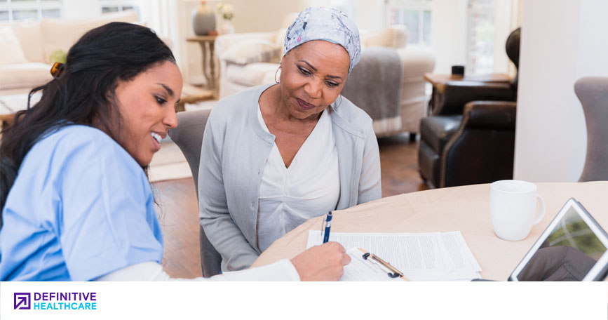 A smiling healthcare worker sits at a table with an elderly patient and helps her fill out paperwork.