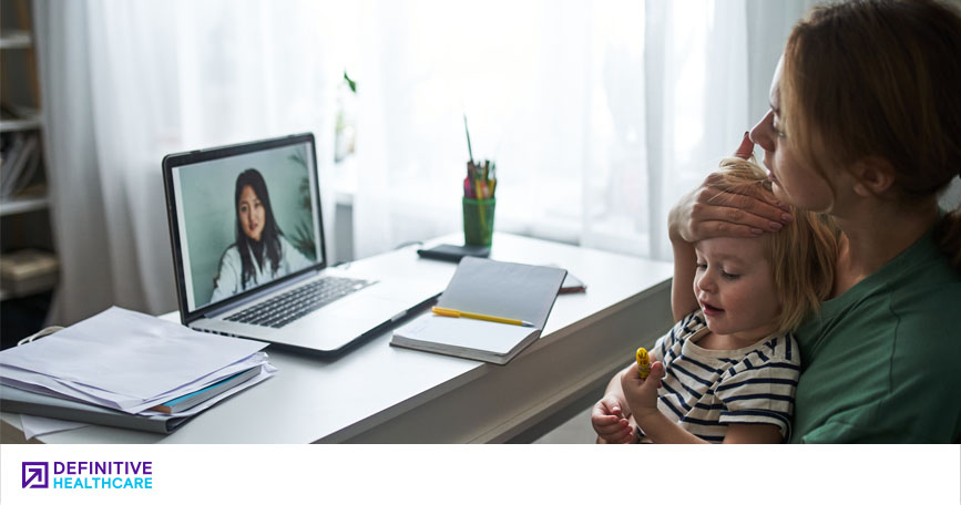 A mother holds her ill child while sitting at a desk and talking with a doctor via telemedicine.