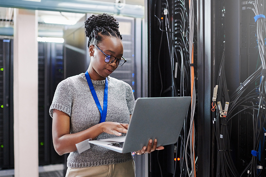 A woman stands in a server room while working on a laptop that she is holding in her arm.