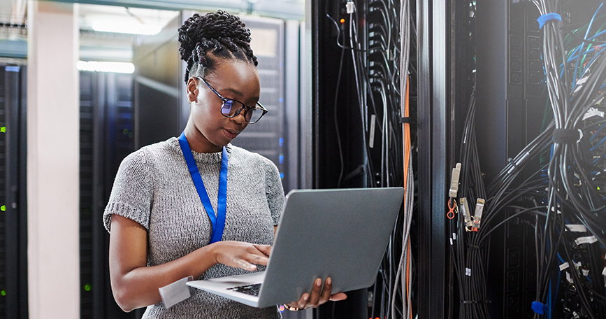 A woman stands in a server room while working on a laptop that she is holding in her arm.