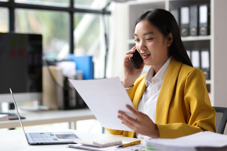 A woman sits at a desk in an office and holds a cellphone up to her ear while looking down at a paper in her other hand.