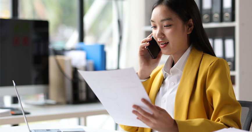 A woman sits at a desk in an office and holds a cellphone up to her ear while looking down at a paper in her other hand.