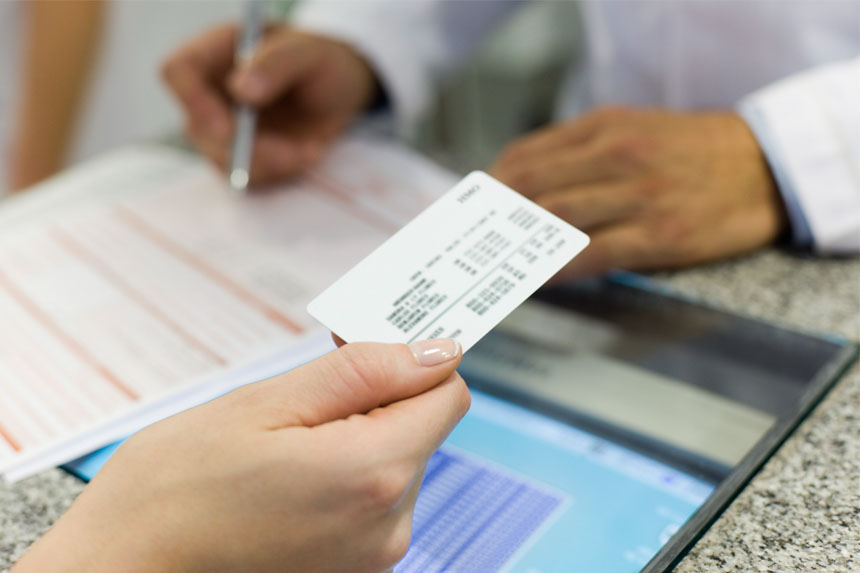 A closeup image of a patient's hand holding an insurance card over a counter while a healthcare professional wearing a lab coat fills out a form.