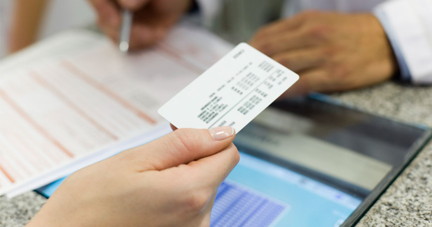 A closeup image of a patient's hand holding an insurance card over a counter while a healthcare professional wearing a lab coat fills out a form.