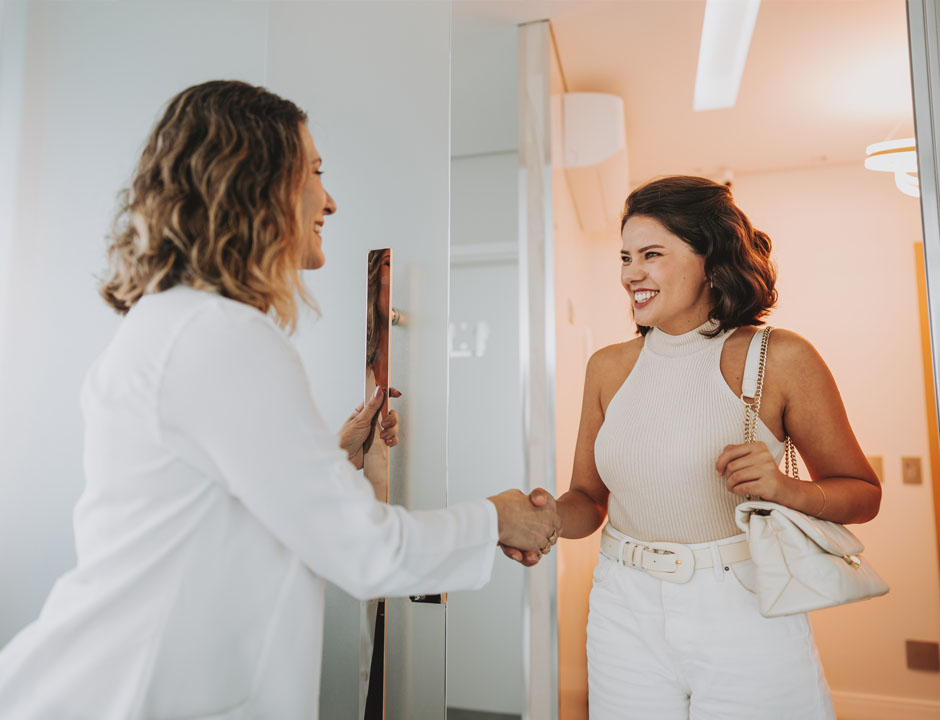 A healthcare professional stands holding a door handle while shaking the hand of a smiling female patient who stands on the other side of the door
