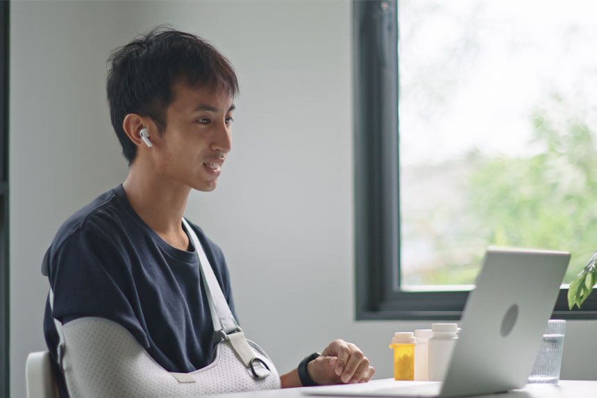 A smiling patient with a cast on his arm sits in front of a laptop computer.