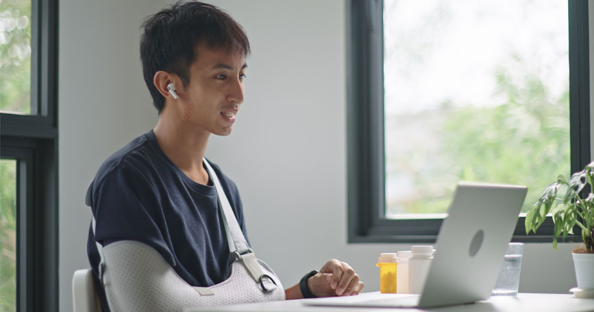 A smiling patient with a cast on his arm sits in front of a laptop computer.