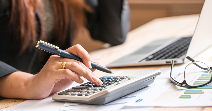 A closeup of a desk and a hand holding a pen as it types on a calculator.
