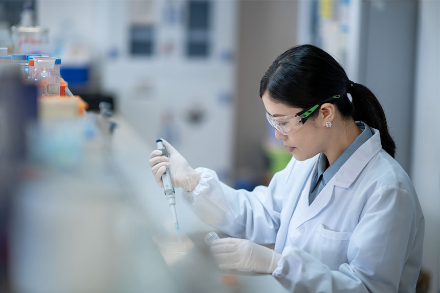 A female researcher in a lab coat and safety glasses uses a handheld instrument in a lab setting.