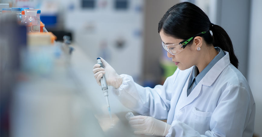 A female researcher in a lab coat and safety glasses uses a handheld instrument in a lab setting.