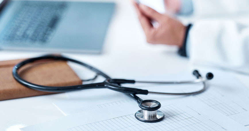 A close-up shot of a physician sitting at a desk. It shows a stethoscope, some papers, a notebook. The physician's arms and laptop keyboard are blurred.