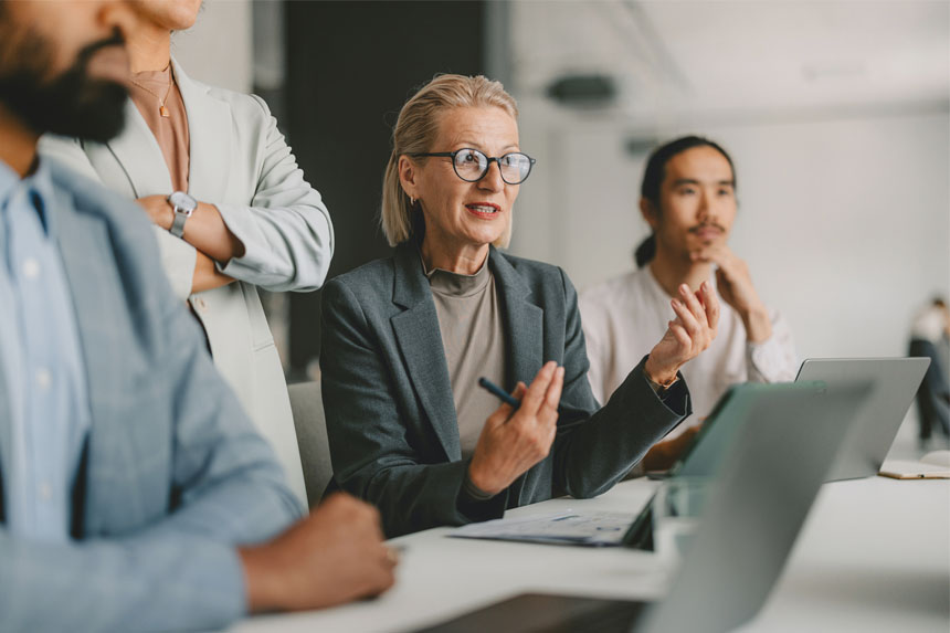 Four professionals dressed in business attire sit and stand at a table having a discussion with someone out of frame.