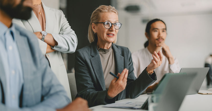 Four professionals dressed in business attire sit and stand at a table having a discussion with someone out of frame.