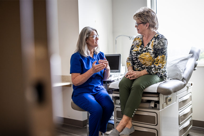 A healthcare professional sits on a stool while talking to a female patient who is sitting on an exam room table next to her.