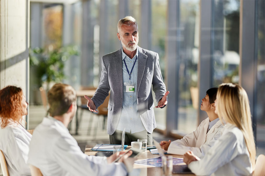 A male dressed in business attire stands at a table while talking to a group of healthcare professionals in white coats.