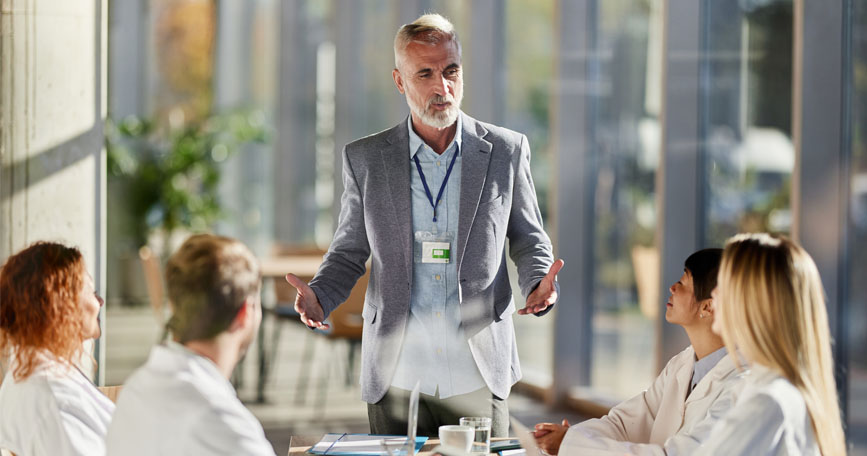 A male dressed in business attire stands at a table while talking to a group of healthcare professionals in white coats.