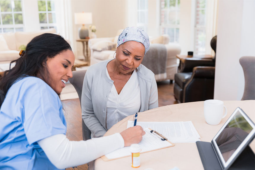 A smiling healthcare worker sits at a table with an elderly patient and helps her fill out paperwork.