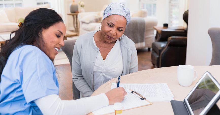 A smiling healthcare worker sits at a table with an elderly patient and helps her fill out paperwork.