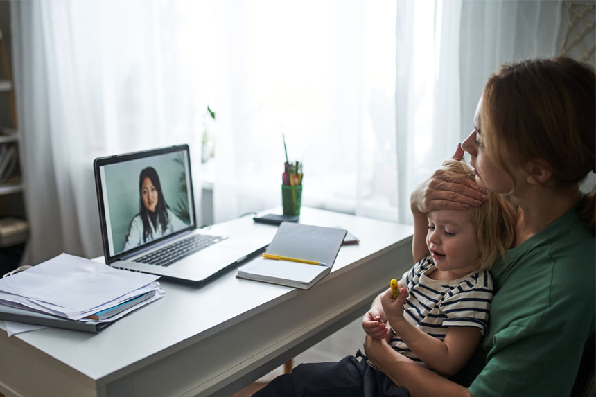 A mother holds her ill child while sitting at a desk and talking with a doctor via telemedicine.