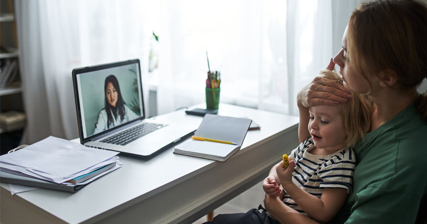 A mother holds her ill child while sitting at a desk and talking with a doctor via telemedicine.