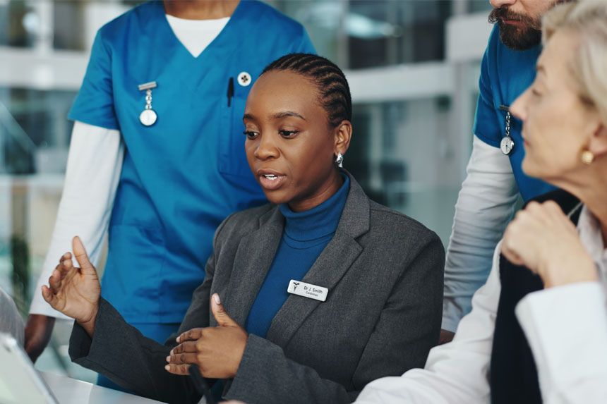 A female doctor dressed in business attire sits while looking down at a screen during a conversation. Two healthcare professionals stand behind her and another professional sits next to her.