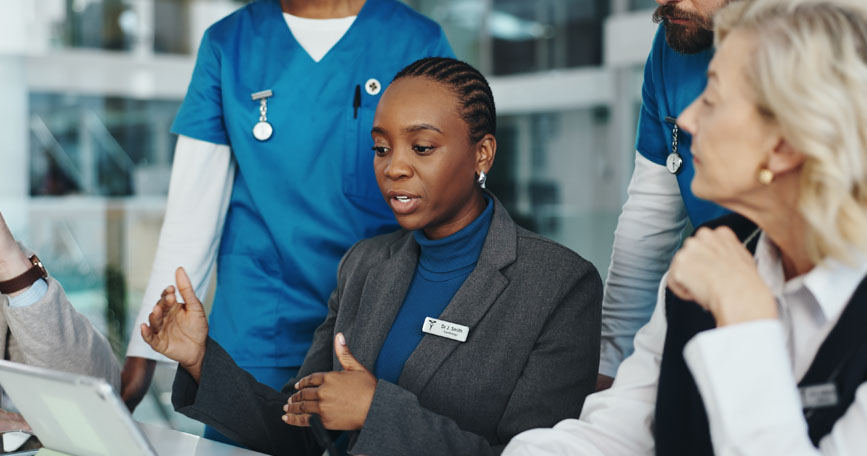 A female doctor dressed in business attire sits while looking down at a screen during a conversation. Two healthcare professionals stand behind her and another professional sits next to her.