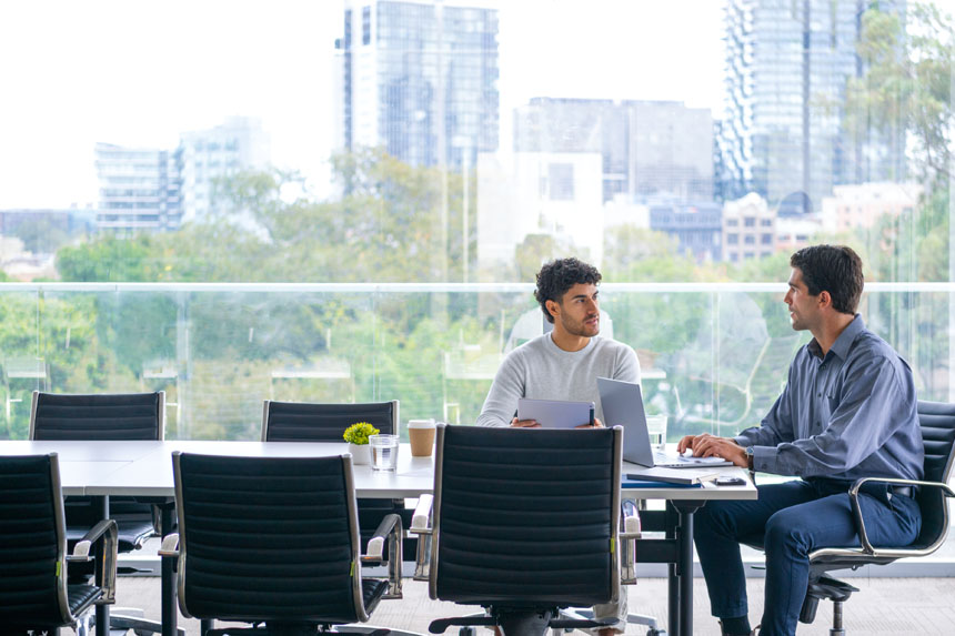 Two males wearing business casual attire sit at a conference room table with a large window in the background having a discussion.