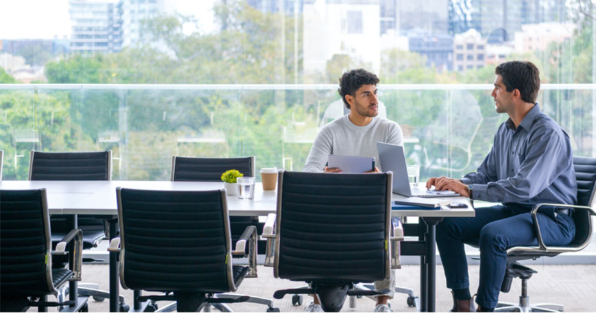 Two males wearing business casual attire sit at a conference room table with a large window in the background having a discussion.