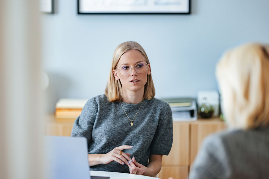 A female healthcare marketing professional sits at a table in front of an open laptop and chats with another team member.
