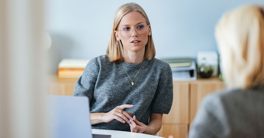 A female healthcare marketing professional sits at a table in front of an open laptop and chats with another team member.
