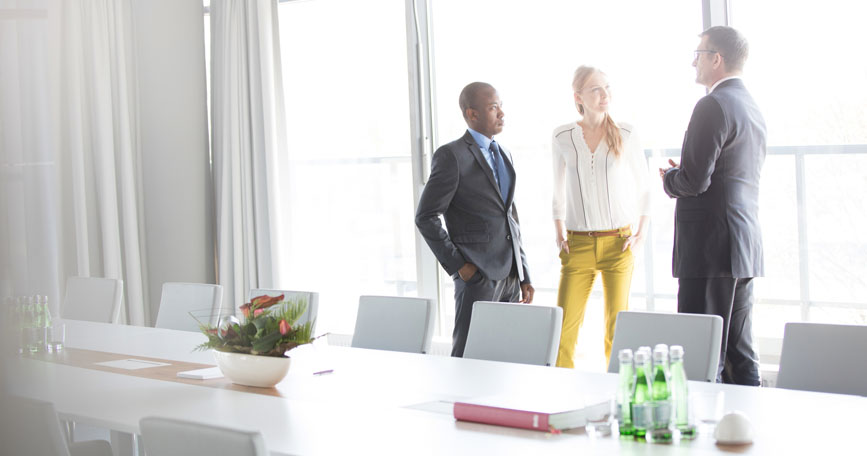 Three professionals in business attire stand in a well-lit conference room having a discussion.