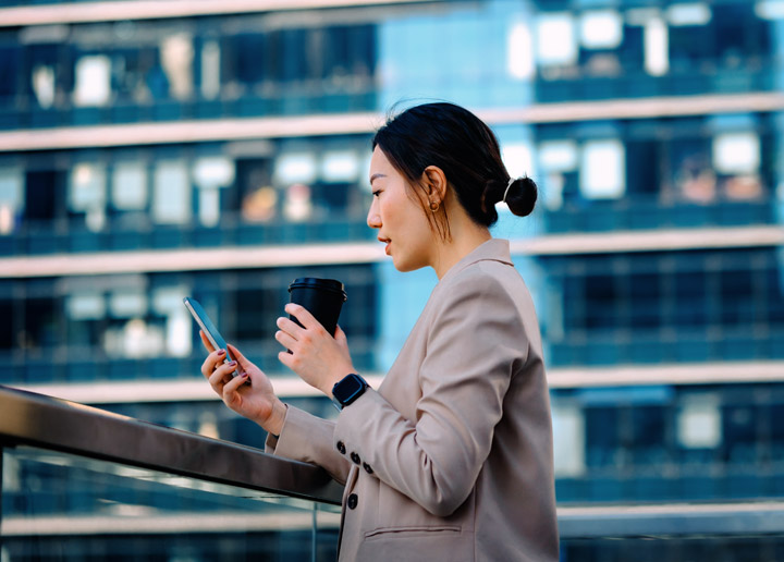 A female dressed in business attire leans against a railing while looking down at a cellphone and holding a cup with a lid.