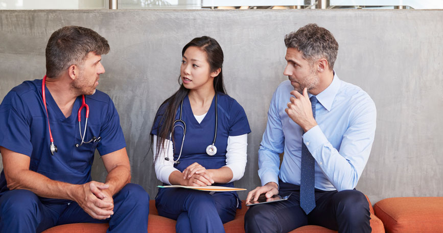 Two healthcare professionals and a male dressed in business attire sit together.