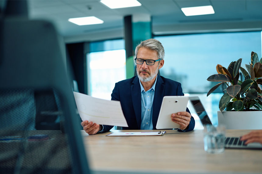 A healthcare operations professional examines a tablet and a printed report while sitting at a desk in a well-lit room. 