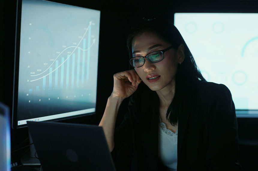 A female analyst sits in a dark office surrounded by computer screens.