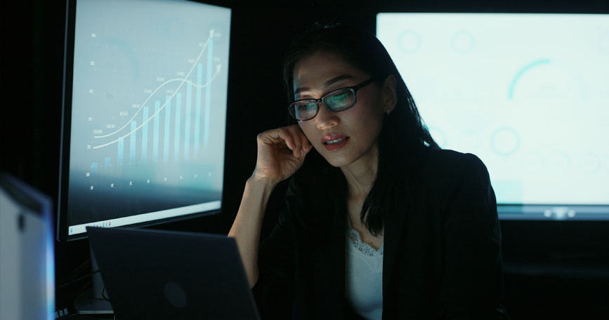 A female analyst sits in a dark office surrounded by computer screens.