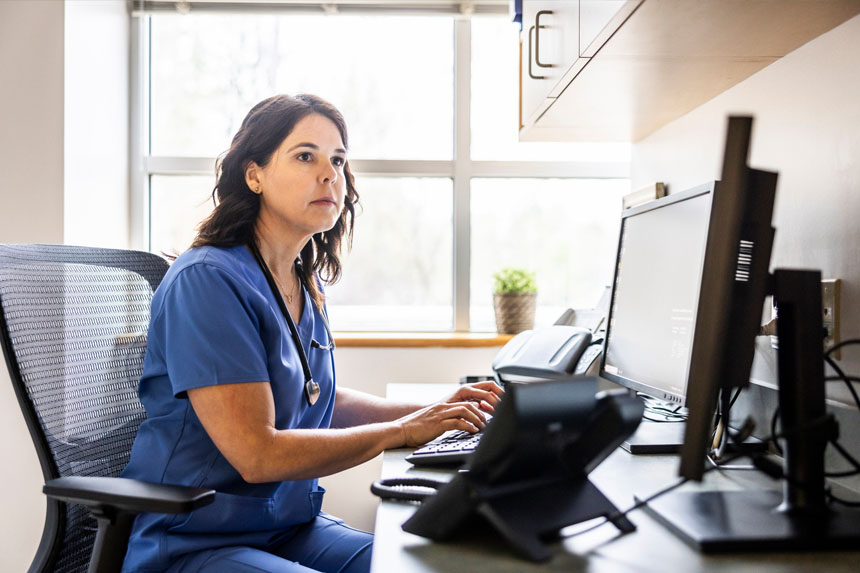 A healthcare professional sits at a desk and types on a keyboard while looking at a monitor screen.
